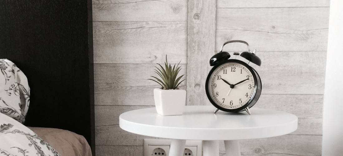 A clock on a white nightstand next to a bed. There is a plant to the left of the clock.